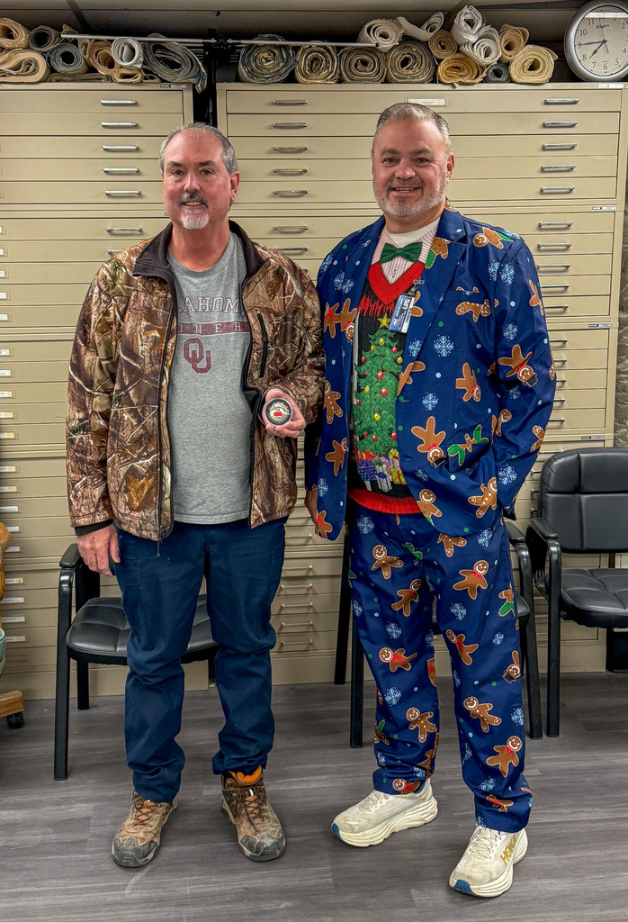 Two men in a room with a wooden floor and storage cabinets behind them. One man wears a camouflage jacket and the other a Christmas sweater.