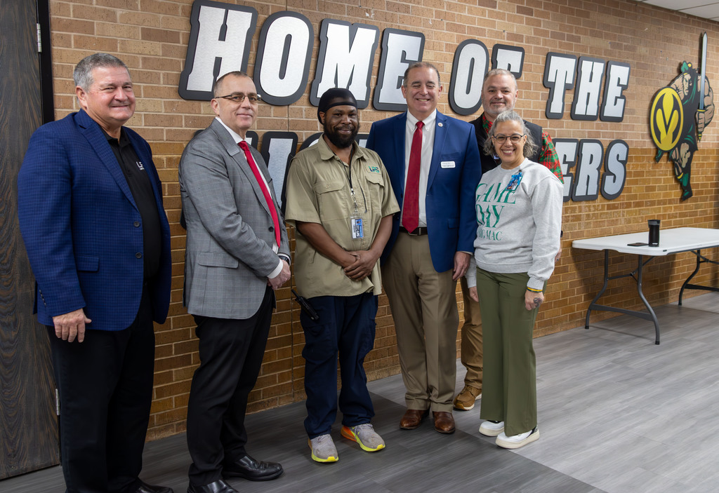 Six people pose in front of a brick wall with "Home of the Warriors" and a logo.