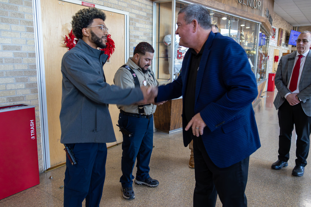Two men shaking hands in a hallway, with a security guard and another man in a suit nearby.