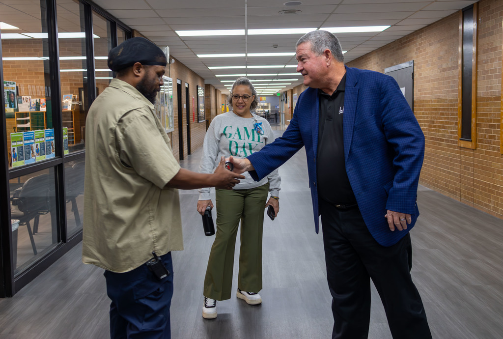 Three people in a school hallway. One man shakes hands with another man in a suit. Behind them, a woman walks.