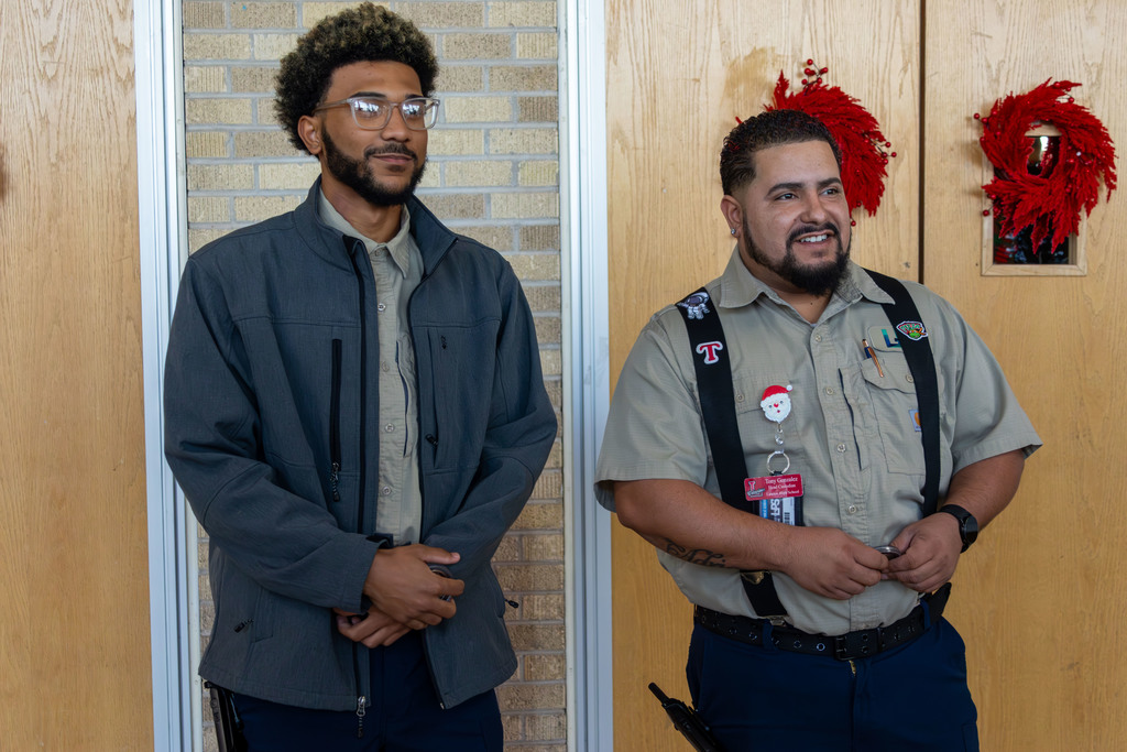 Two men stand before a brick wall, one in a jacket and the other in a uniform with suspenders.