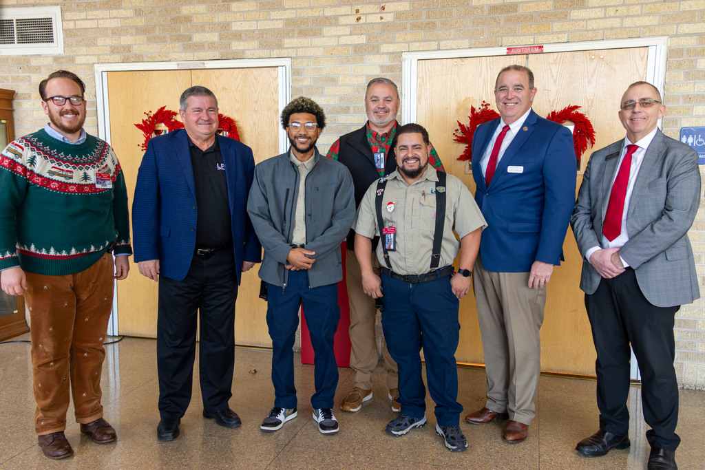 A group of men dressed in winter attire stand in a line in a hallway. One man wears a Christmas sweater.