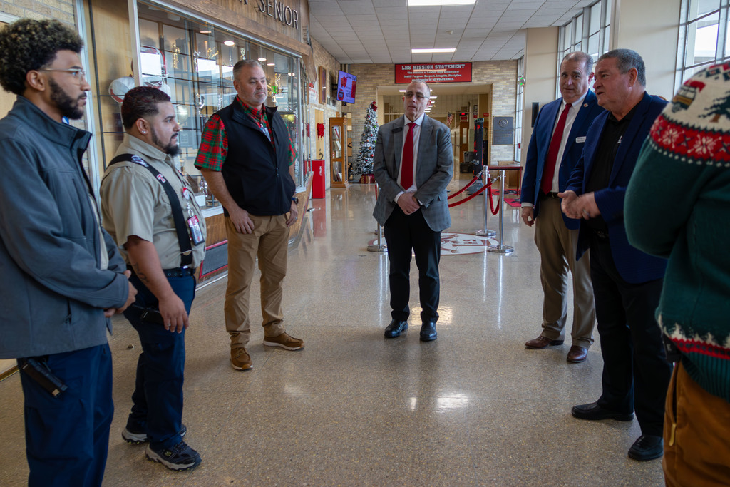 Men in various attires, including suits, vests, and shirts, stand in a hallway, discussing. A festive sweater, monitors, and a rope barrier are present.
