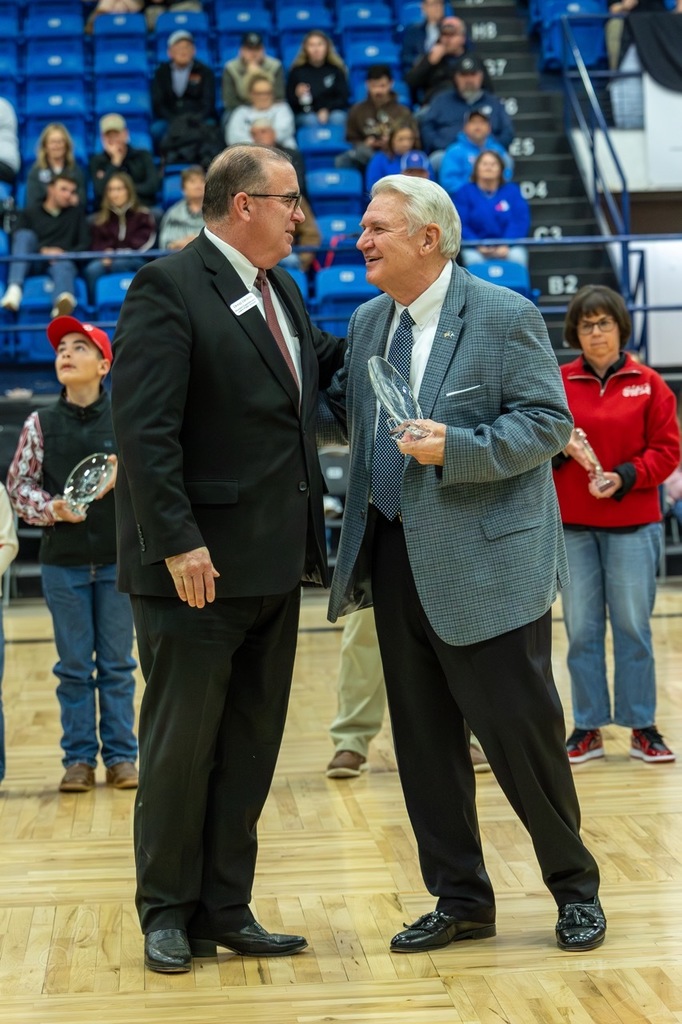 Two men in suits stand on a basketball court, one holding an award, while spectators watch from blue bleachers.