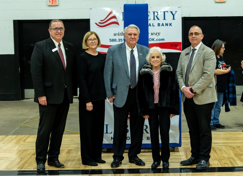 A group of five people stand on a basketball court. Three men and two women in suits. Behind them, a sign with the word "ERTY" and an American flag.