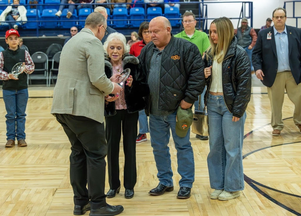 A group of people stand on a basketball court, two men exchanging an award. One woman holds a trophy.
