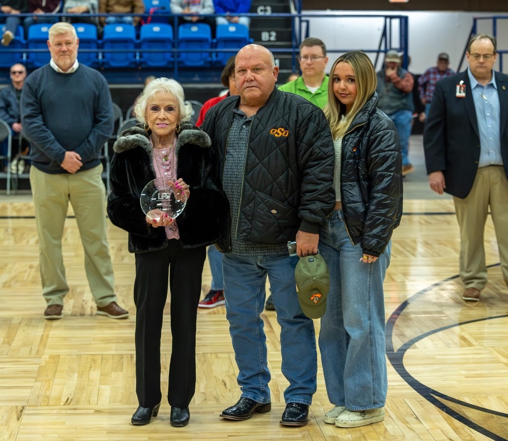 Group of people on a basketball court, one holding an award, surrounded by others. Empty blue bleachers in the background.