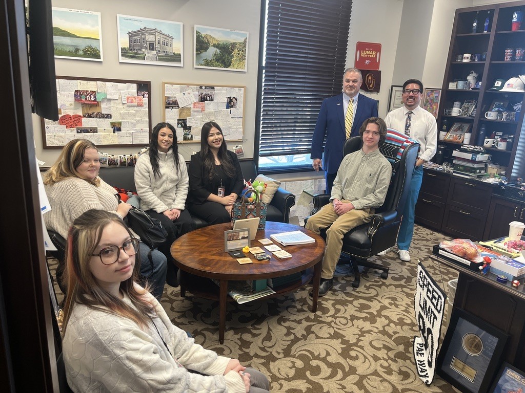 Group of people sit in chairs in a room with a wooden desk and bookshelves.