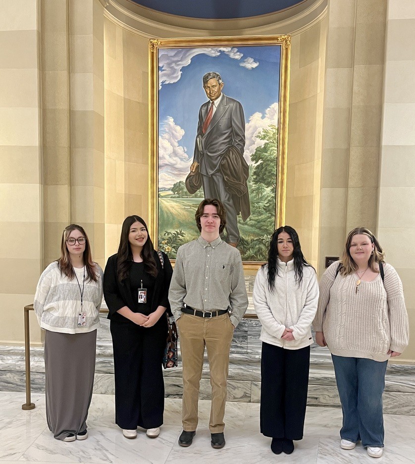Five individuals pose in front of a large portrait of a man in formal wear.