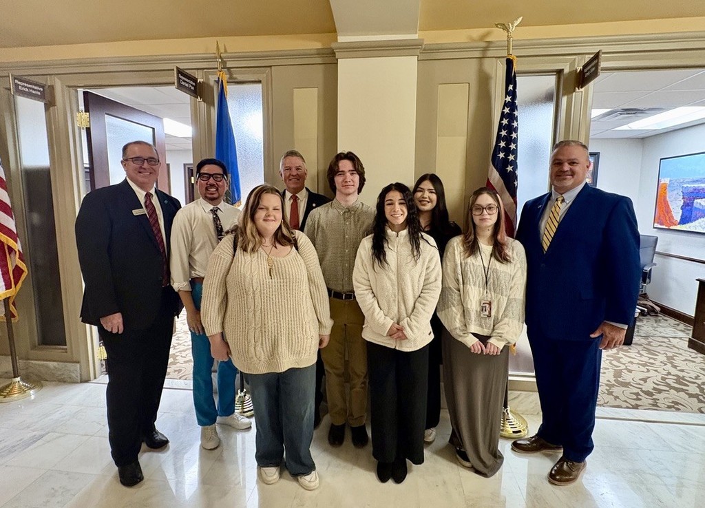Group of people in suits, ties, and casual clothing standing together in a hallway with flags.