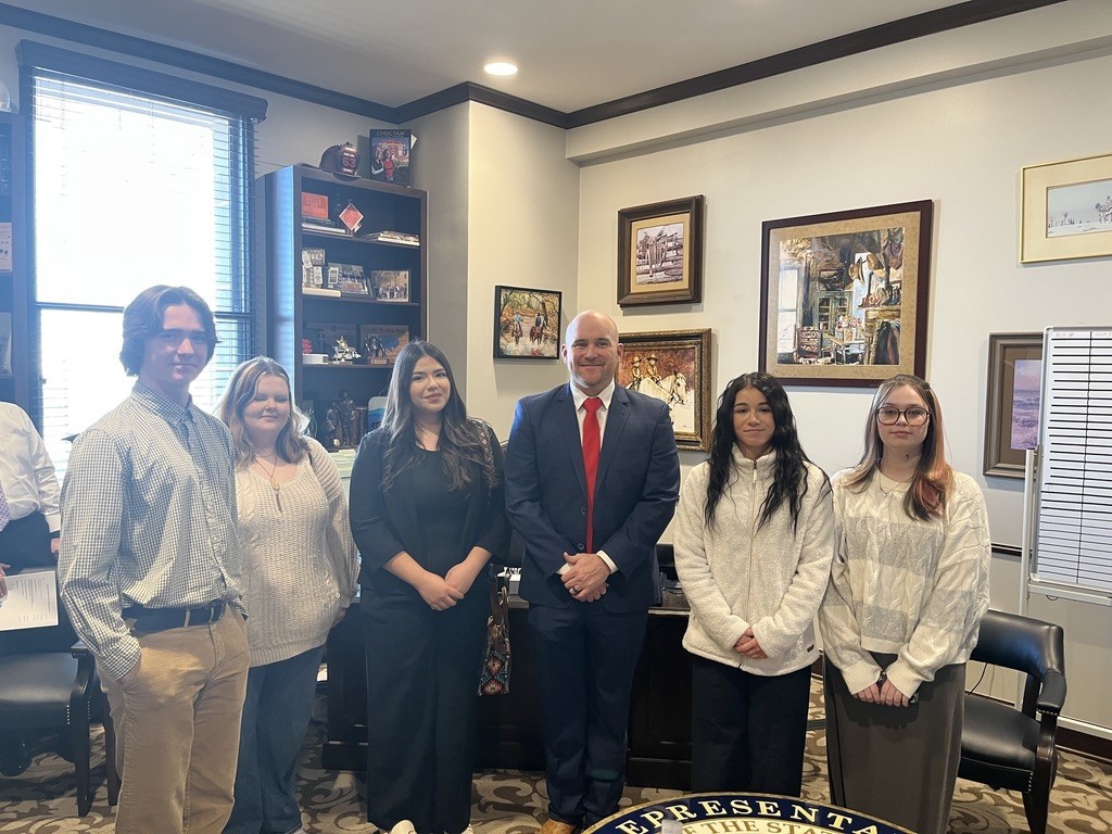Group of people, including a man in a suit and tie, standing in a room with a carpeted floor.