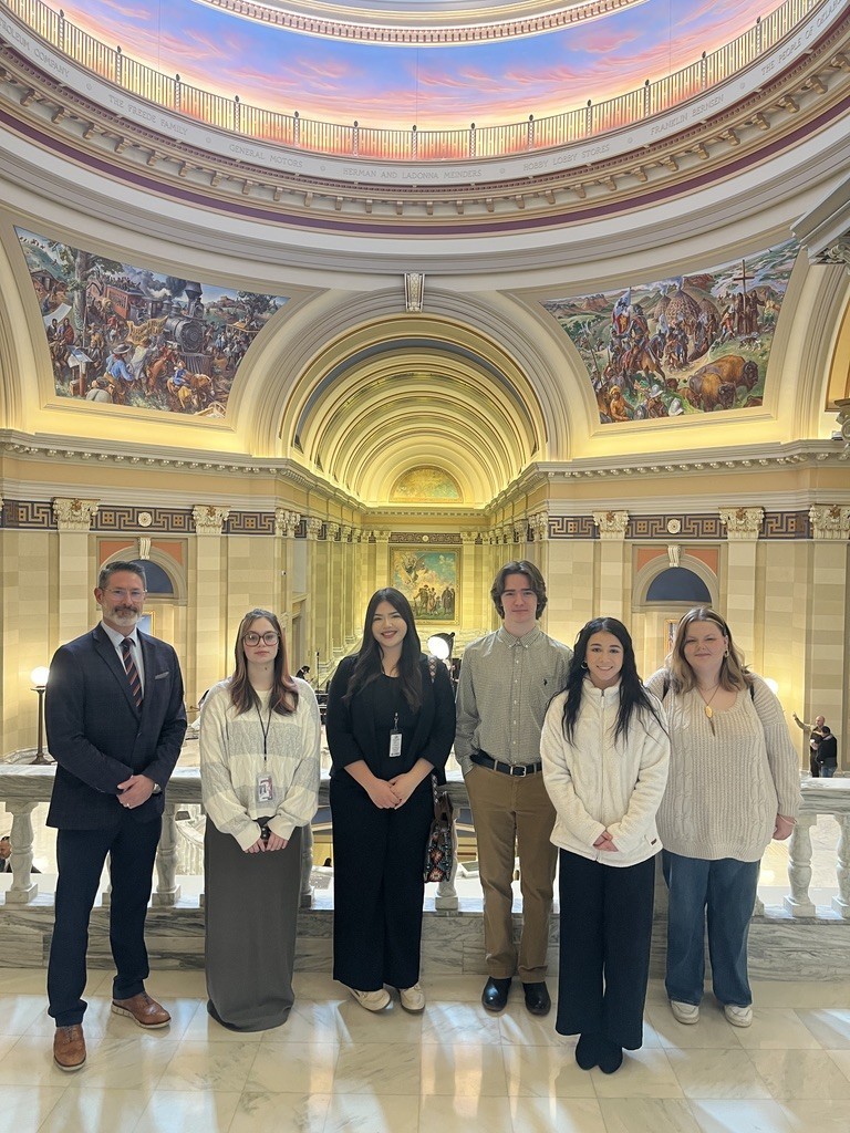 A group of people stands in a grand room with high ceilings and arched windows. They wear casual and formal clothing.