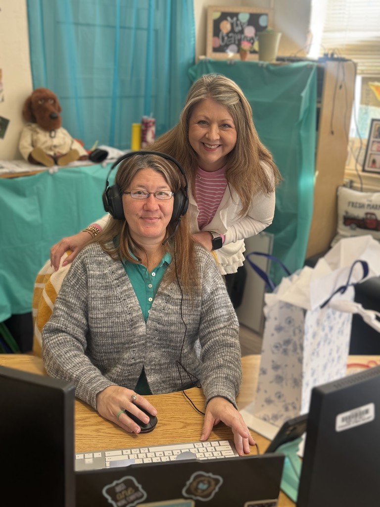 Two women in a room with a computer desk. One woman wears headphones and sits. Another woman stands behind her.
