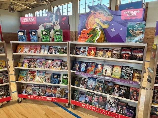 A library with a dinosaur display, three shelves with colorful book covers, and a wooden floor.