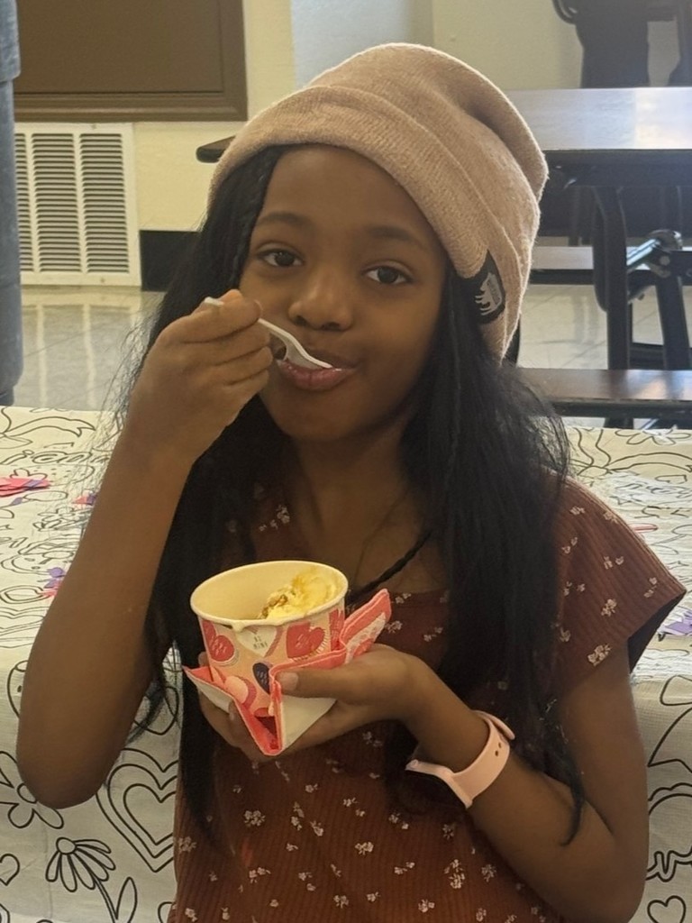 A young girl eats ice cream from a cup while seated at a table. She wears a pink beanie and a brown top with a floral design.