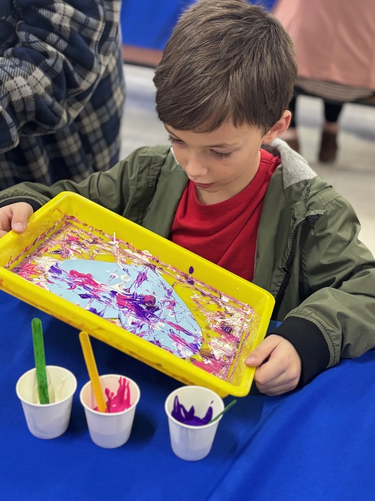 A young boy wearing a green jacket is seated, painting with paint on a yellow tray.