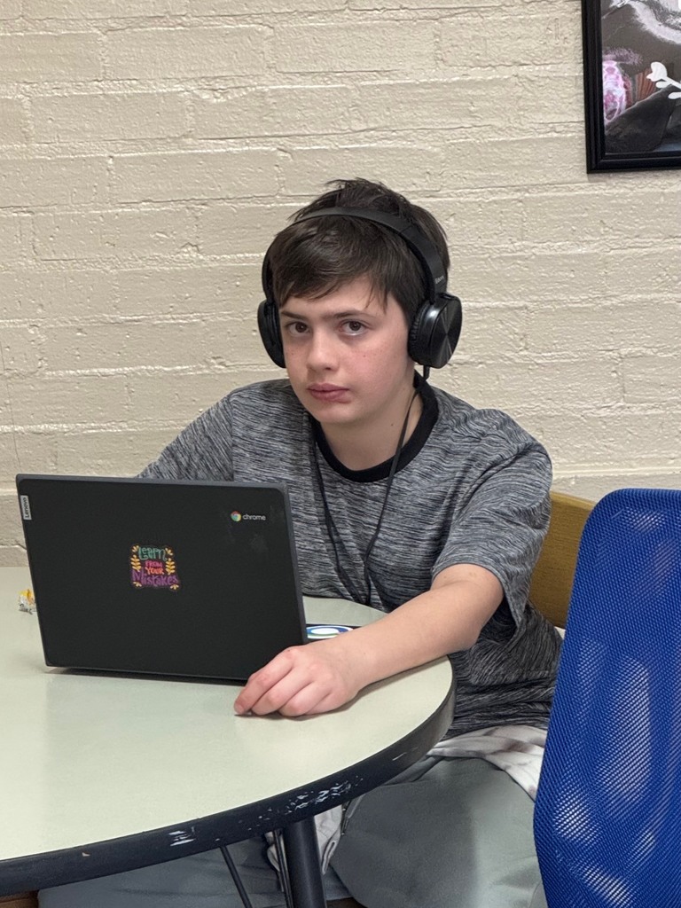 A boy wearing headphones and a gray shirt sits at a table with a laptop.