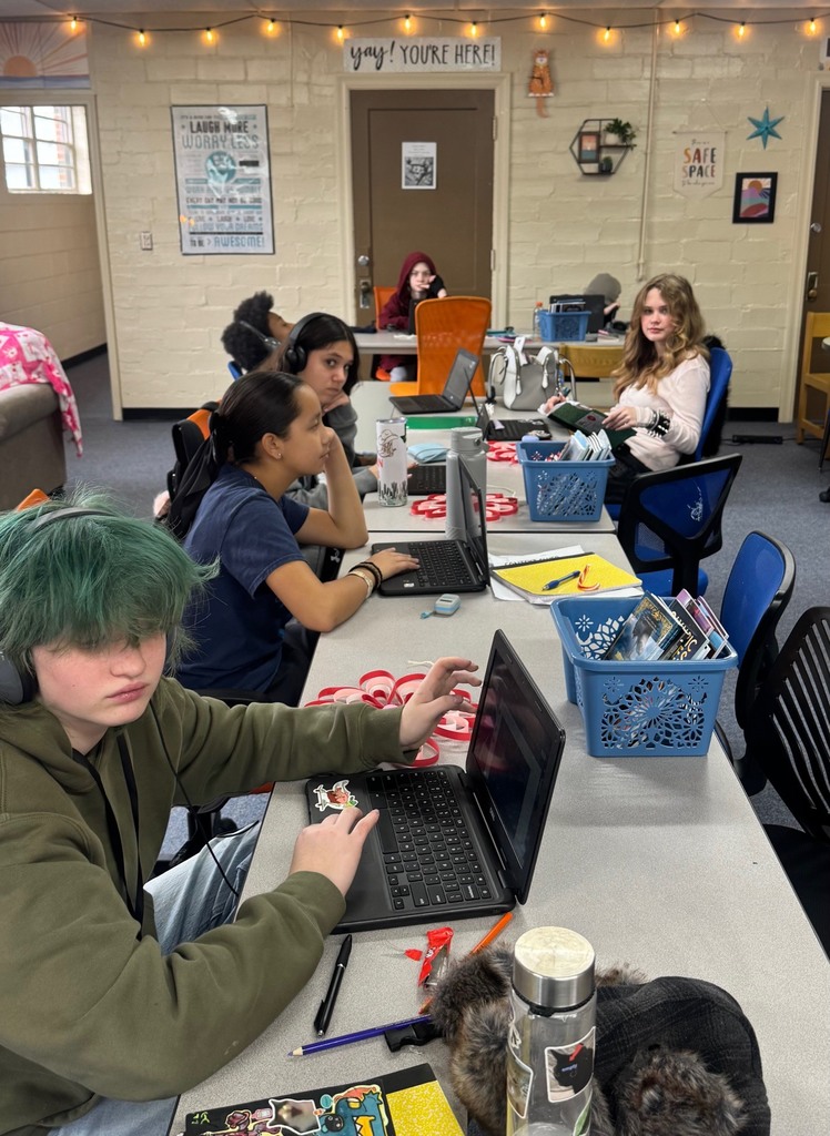 A group of people sitting around a table with laptops, including one wearing green hair and headphones.