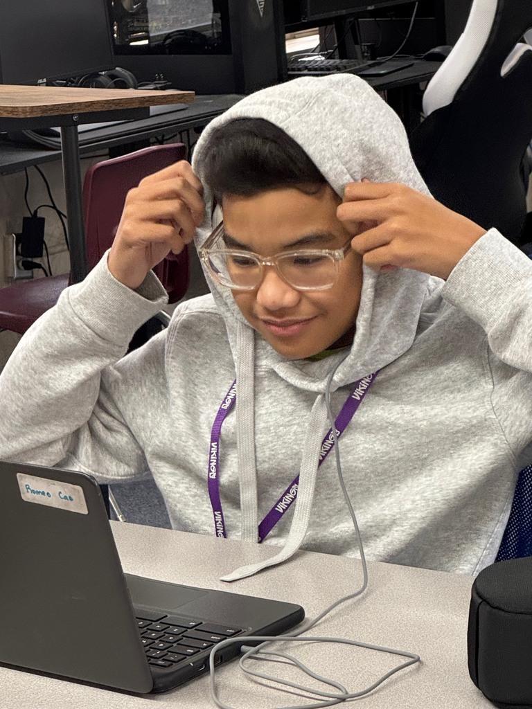 A person in a gray hoodie sits at a desk, using a laptop. They wear glasses and have headphones.