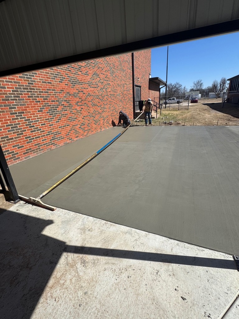 Two workers are smoothing cement on a driveway using a tool, with a brick building in the background.