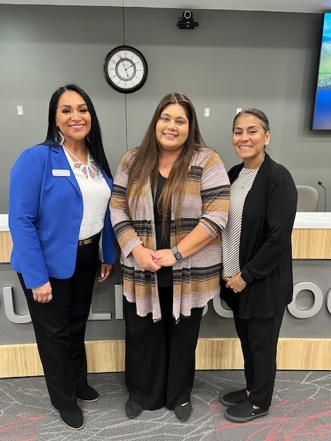 Three women stand in front of a reception desk. They are wearing suits and are smiling. A clock is on the wall behind them.
