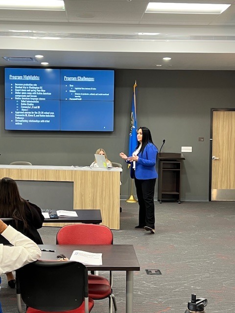 Woman standing at a podium in front of a screen, speaking. Two tables with chairs and a flag.