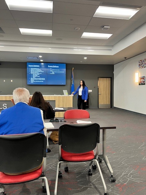 A woman in a blue jacket stands at a podium in a meeting room. Two people sit at a table.