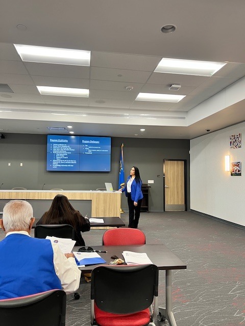 A woman stands in front of a screen, likely giving a presentation. An audience sits in chairs, facing her.