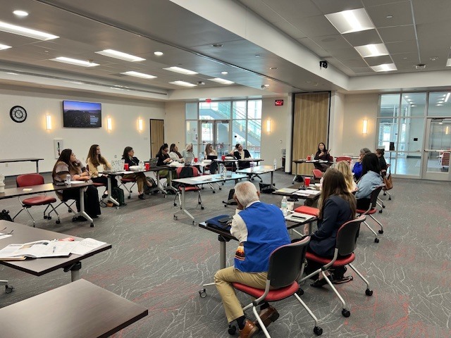 Room with tables and chairs, people sitting and working, papers and notebooks on tables, ceiling lights, glass doors.