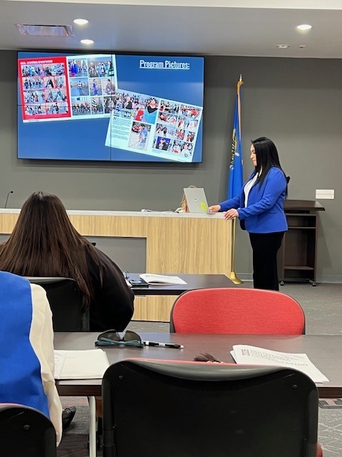 A woman in a blue blazer stands at a podium, presenting to an audience. A screen behind her displays a collage of photos.