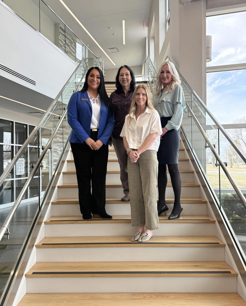 Five women stand on a flight of stairs, posing for a photo. The stairs are made of wood.