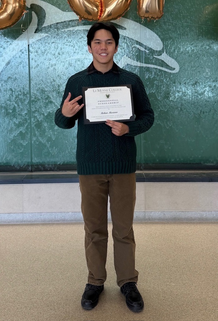 Man stands in front of a blue wall, holding a certificate. Behind him are three gold balloons.