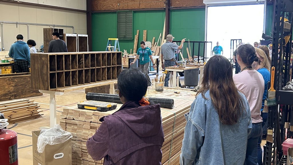 People are gathered in a workshop with wooden shelves and planks. Some are observing the area.