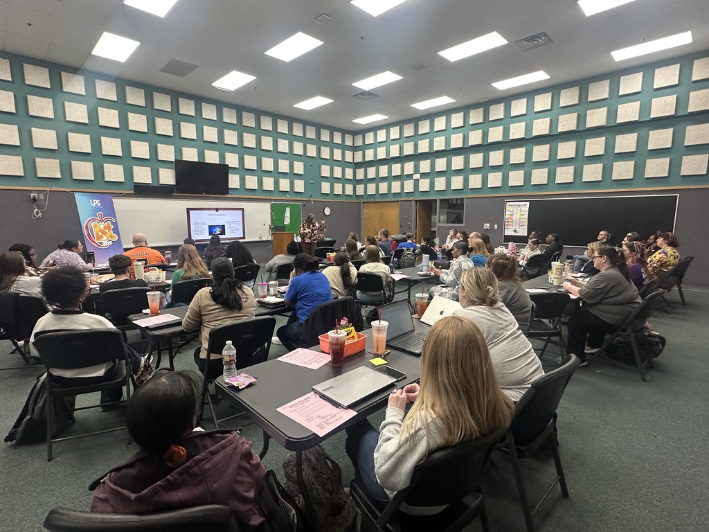 A room with square tiles on the wall and ceiling lights. People sit in chairs at tables.