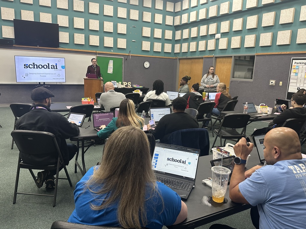 A classroom with people seated at tables using laptops, one man giving a speech at a podium.