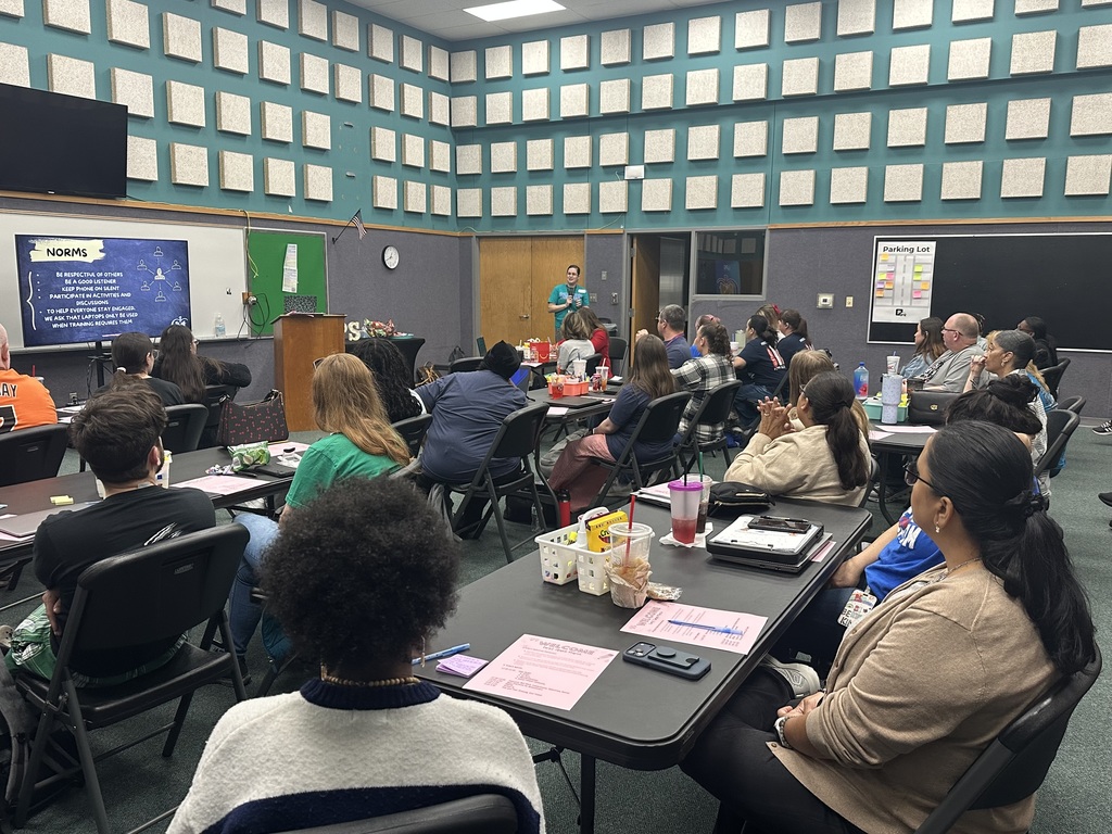 A classroom with a teacher at a podium. Attendees sit at tables, listening. A projector screen is on the wall.