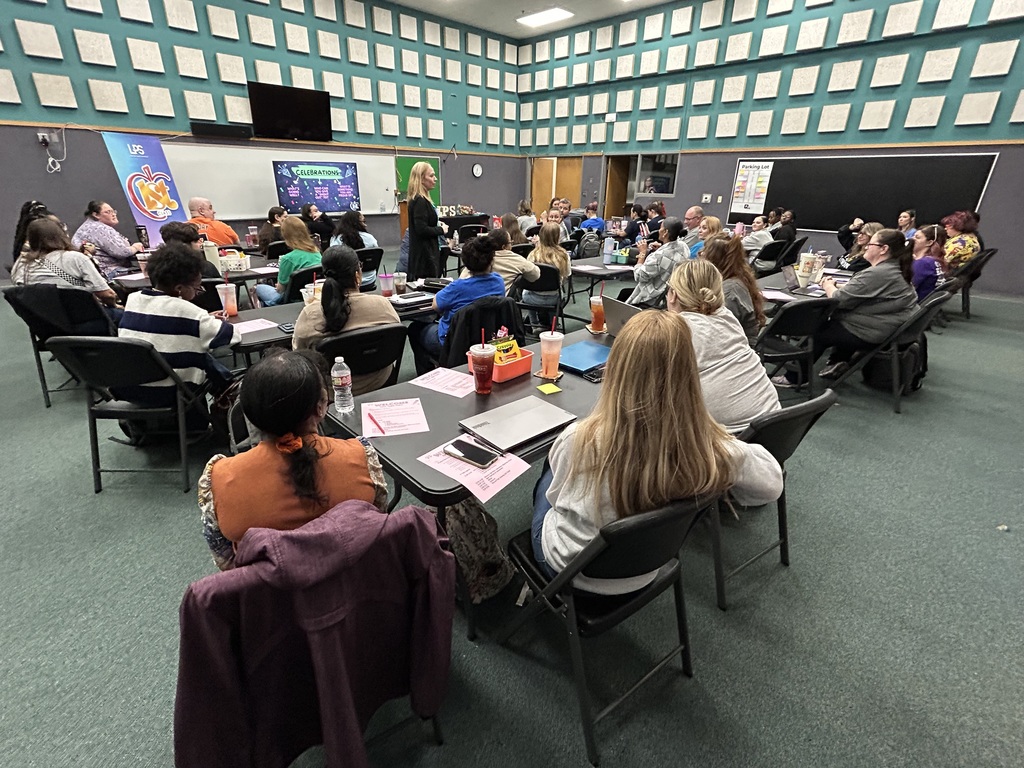 A group of people sit at tables in a classroom with a green-patterned wall. One person stands at a front desk.
