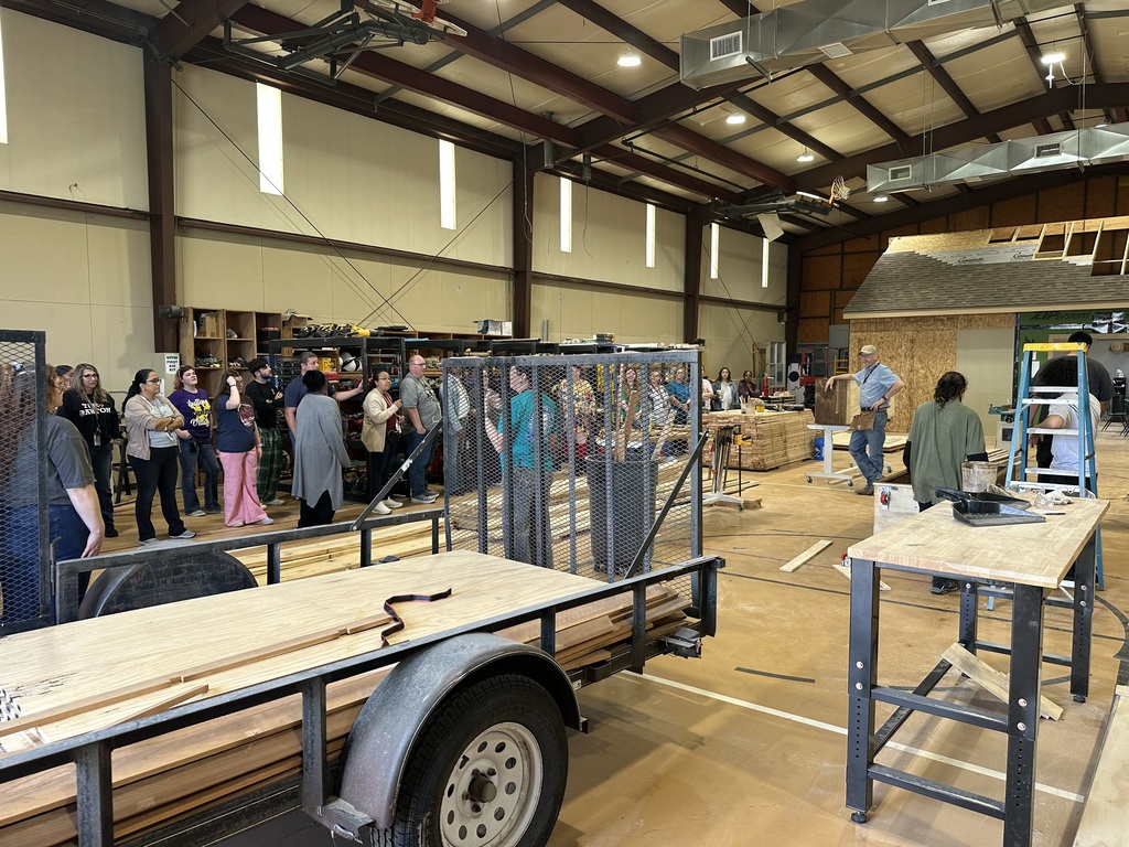Several people gather in a large workshop, surrounded by wooden materials and equipment. A trailer is parked on the left.