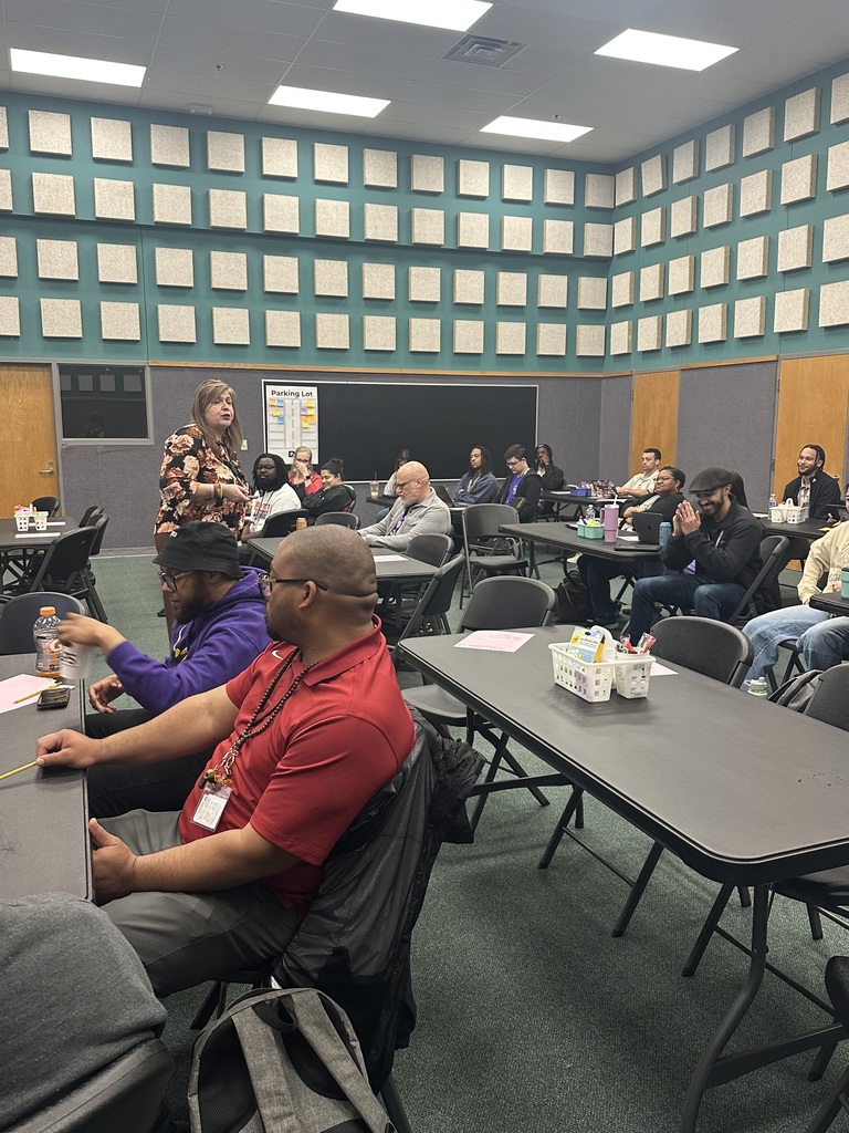 Room with tables and chairs. A woman stands at the front, and people sit. Green walls with squares and overhead lights.