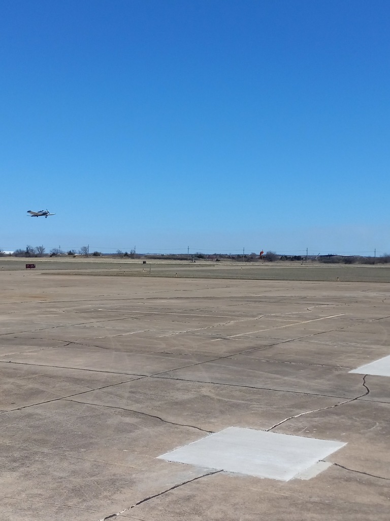 An empty runway with two white squares on it. An airplane flies above it. Background shows a clear blue sky.