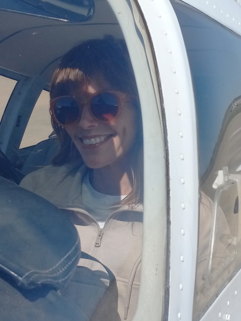 A woman wearing sunglasses and a jacket smiles in an airplane cockpit. The background shows a blurred pilot's seat and a window.