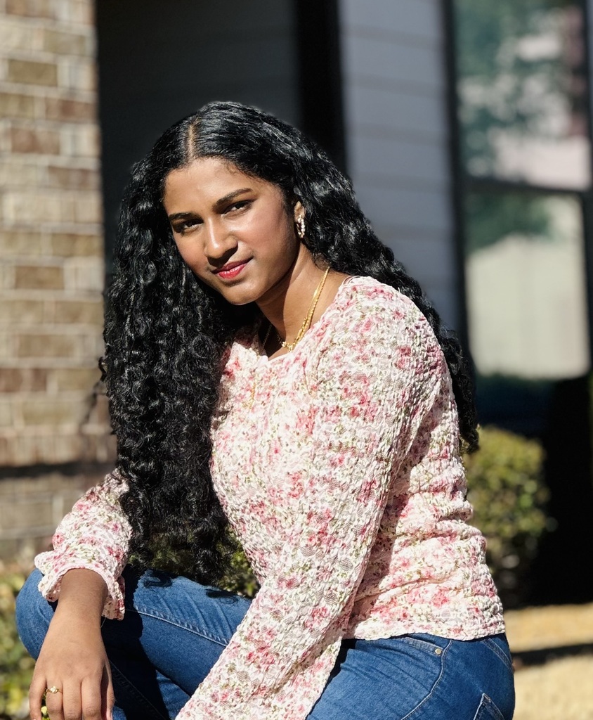 Woman with long, curly black hair in a pink floral top and blue jeans kneeling outdoors.