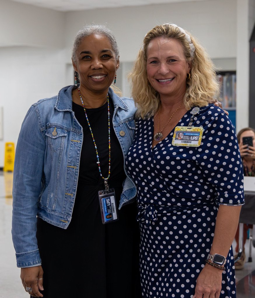 Two women, one with a denim jacket, the other in a blue polka-dot dress, stand next to each other in an indoor space.
