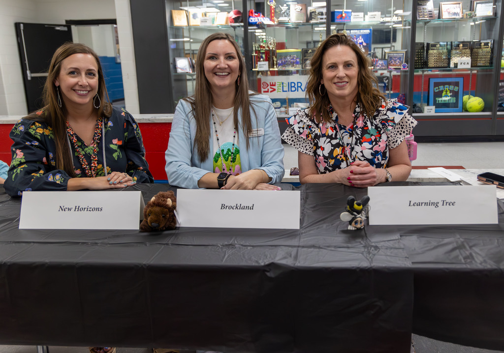 Three women seated at a table with name placards. Behind them, a room with shelves and items.