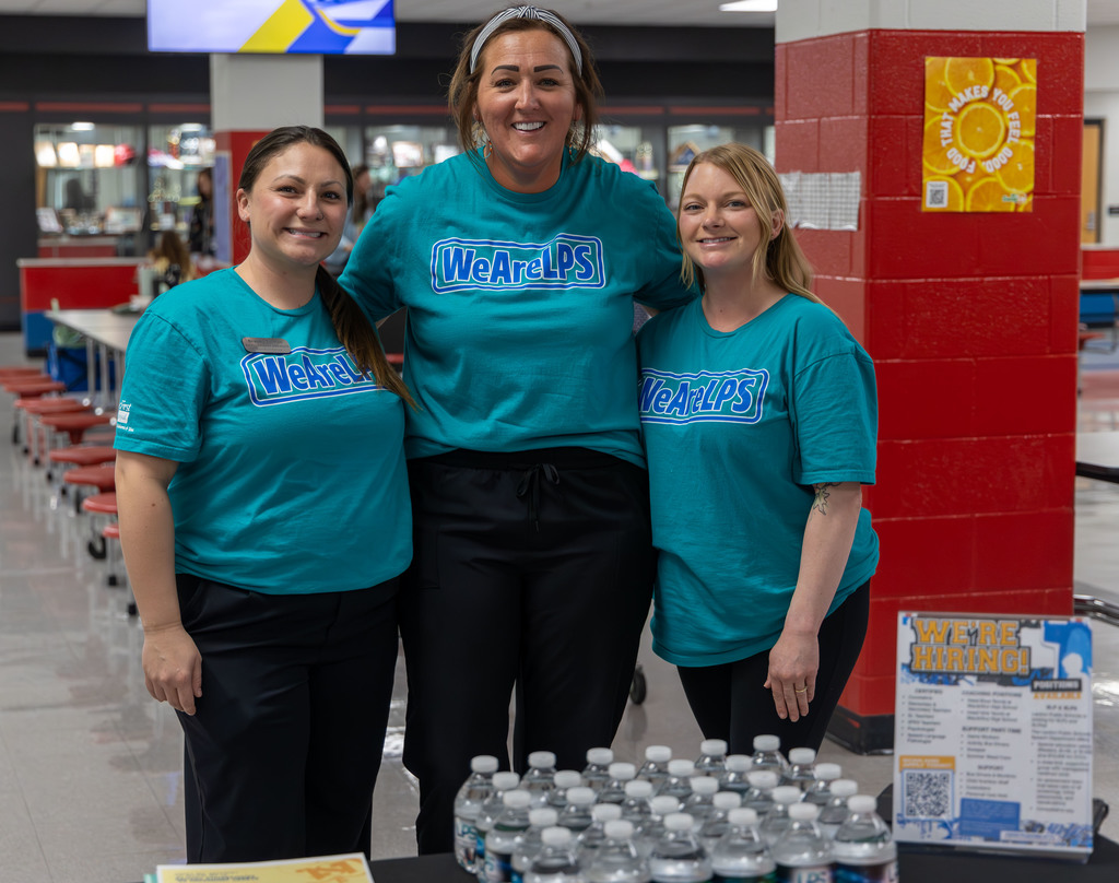 Three women in teal shirts stand at a table with water bottles. They are smiling and facing the camera. Behind them, a red column displays a yellow poster.