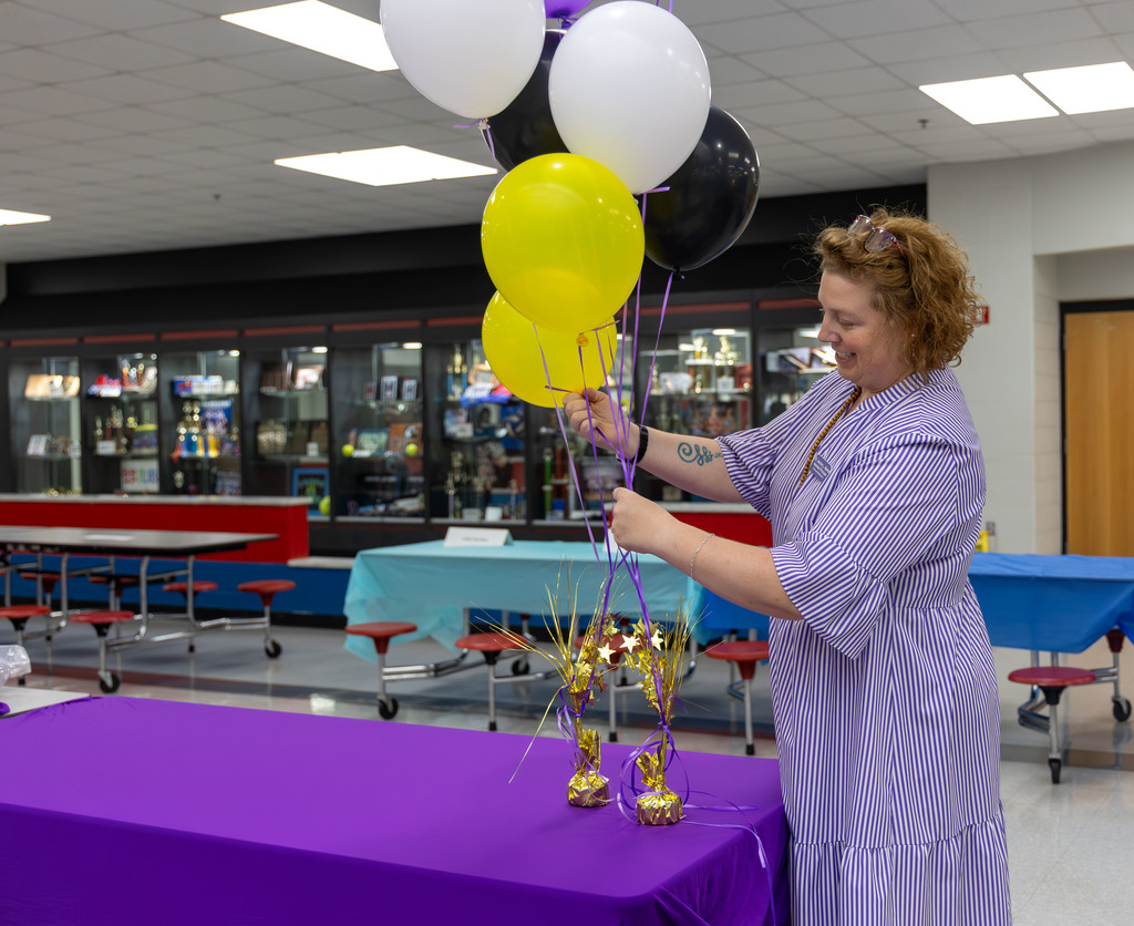 A woman in a purple dress holding a bundle of black, white, and yellow balloons. She is in a room with tables.