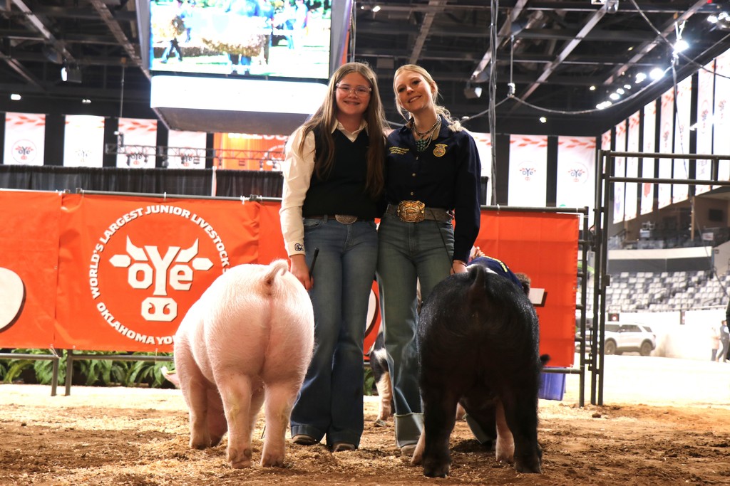Two women stand next to a white pig and a black pig in an arena with a red banner.