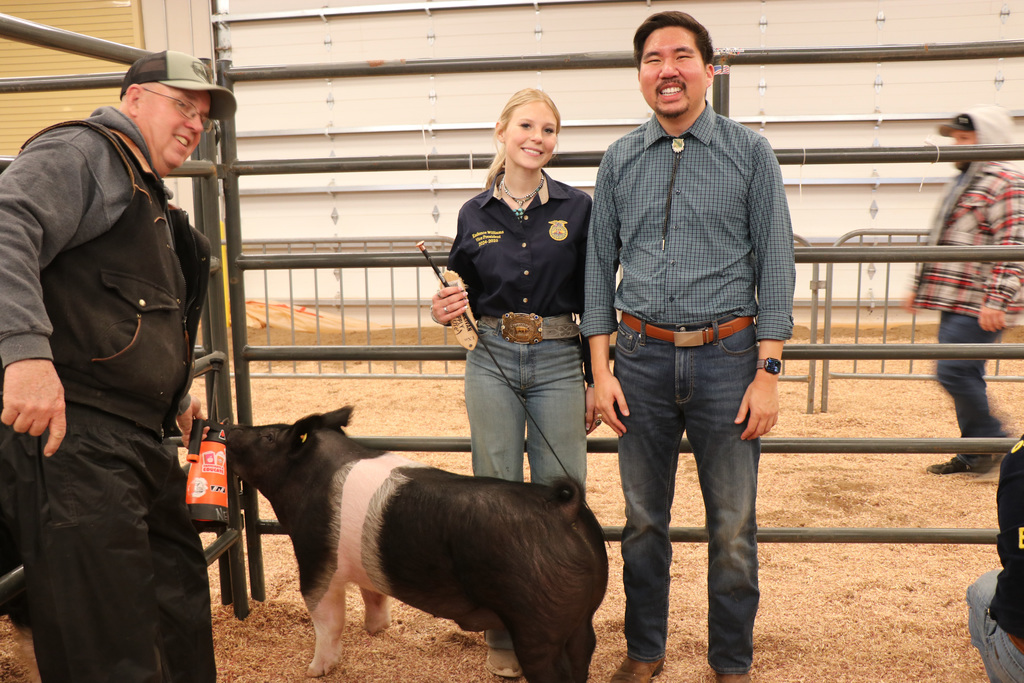 Three people stand behind a metal fence. A small black and white pig stands in front. Background shows a dirt floor and more metal fencing.