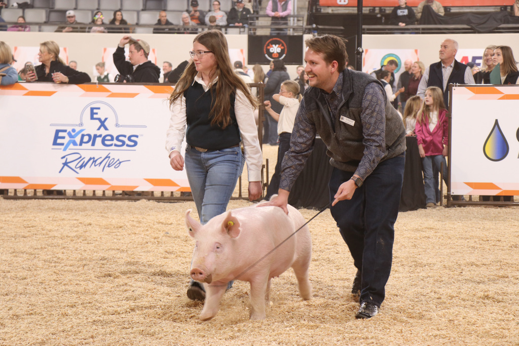 A man and woman with a pig on a leash in an arena. Behind them, people watch from the stands.