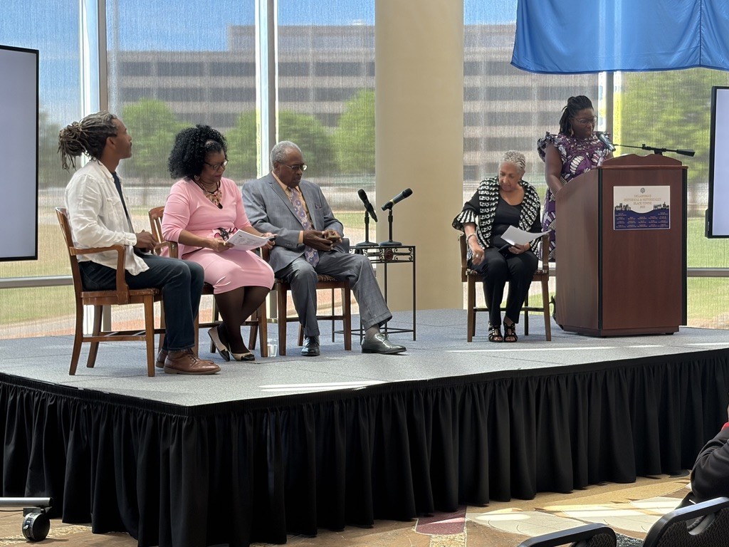 A group of five people on stage, each seated in a chair. A podium with a microphone is present.